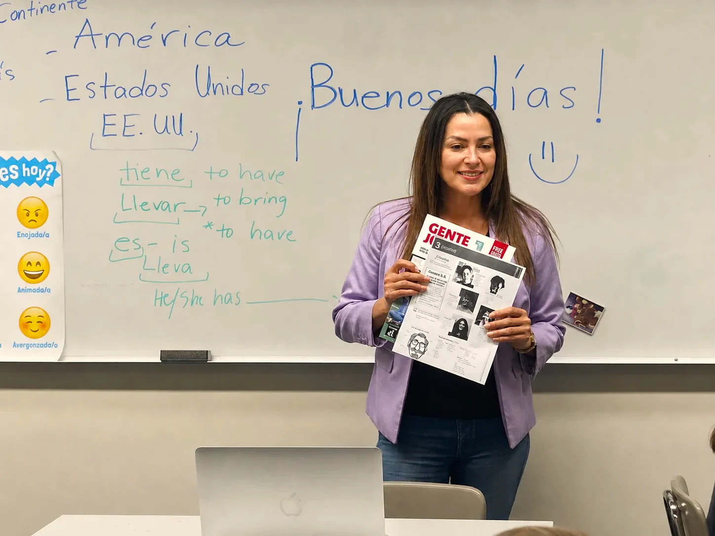 Spanish instructor holding papers in front of a whiteboard with vocabulary notes and “Buenos días” written on it.