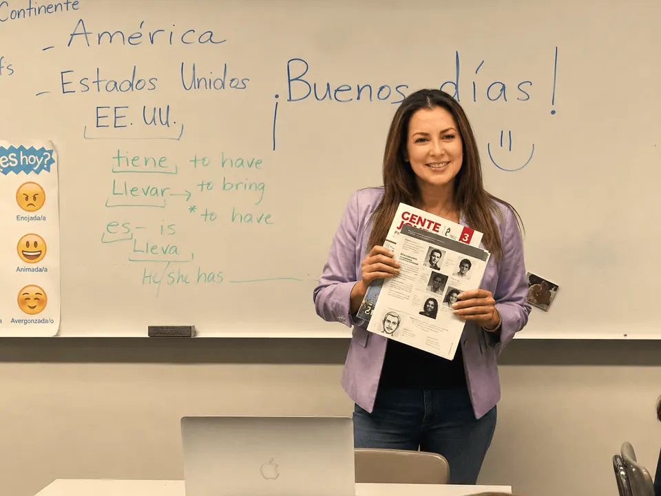 Spanish instructor holding papers in front of a whiteboard with vocabulary notes and “Buenos días” written on it.